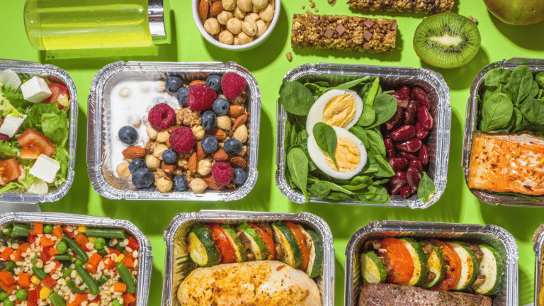 This image showcases an assortment of meals from the best prepared meal delivery service, arranged neatly in aluminum containers on a bright green background. The selection includes a well-balanced salad, a nutritious yogurt bowl with fresh berries and nuts, protein-packed dishes with hard-boiled eggs and spinach, and a succulent salmon fillet. Also visible are a colourful medley of steamed vegetables, a hearty chicken and vegetable dish, and sides of red kidney beans. The ensemble is complemented by a bottle of green juice, a wholesome granola bar, and a ripe kiwi, demonstrating the variety and quality offered by top-tier meal delivery services.